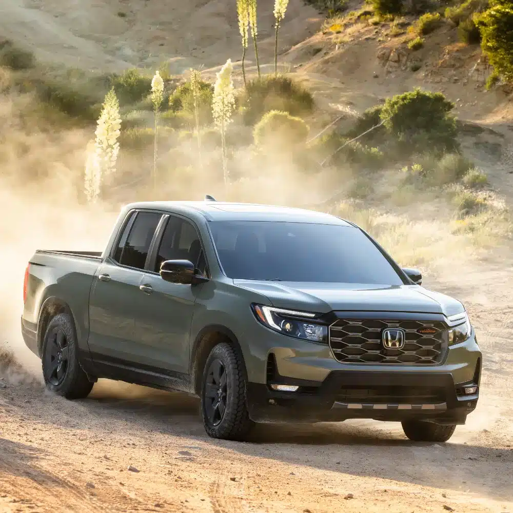 A silver Honda Ridgeline pickup truck confidently drives through a muddy off-road trail.